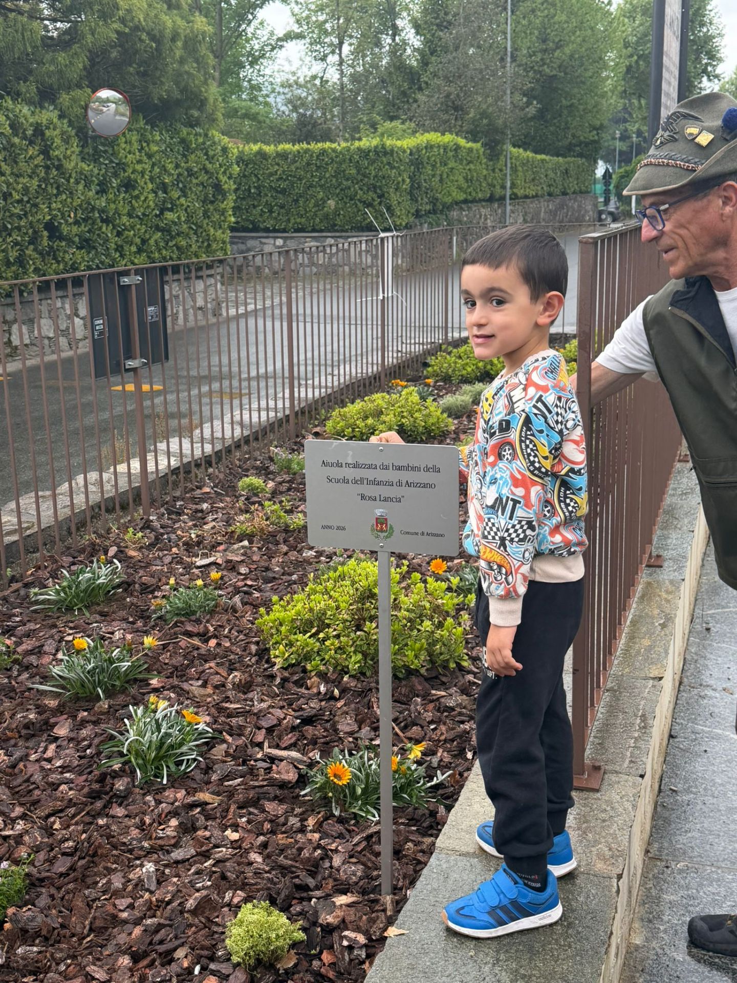 Bambino davanti a targa nell'aiuola con a fianco alpino